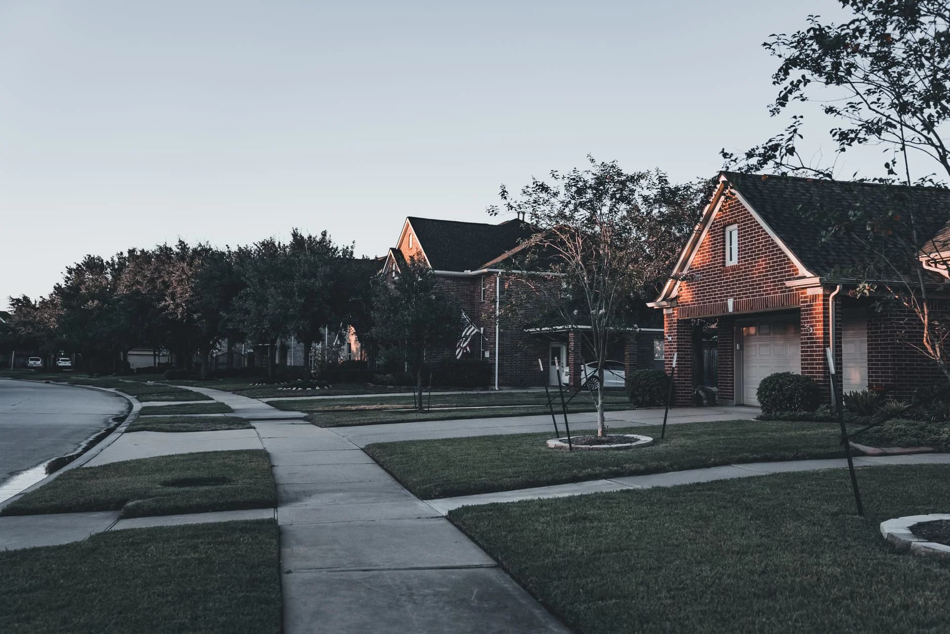A brick house and landscaped front yards along an established residential street at dusk
